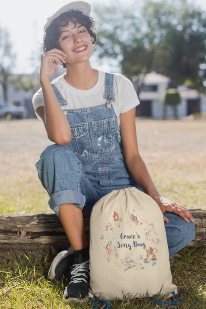 Image of woman holding a cream coloured cotton drawstring bag. Printed on the bag is some colourful illustrations from nursery rhymes, humpty dumpty, little bo peep, a clock, a boot, a goos, a cat and the fiddle and a cow and a moon. The watercolour illustrations are arranged in a wreath shape with the words 'Grace's son bang' written in blue in the middle.