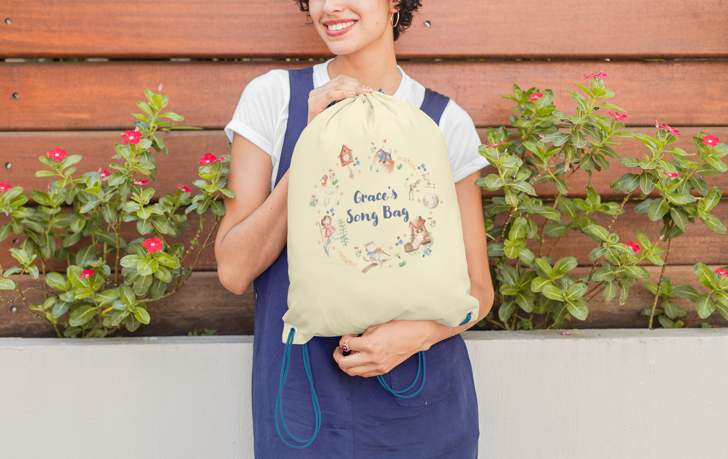 Image of woman holding a cream coloured cotton drawstring bag. Printed on the bag is some colourful illustrations from nursery rhymes, humpty dumpty, little bo peep, a clock, a boot, a goos, a cat and the fiddle and a cow and a moon. The watercolour illustrations are arranged in a wreath shape with the words 'Grace's son bang' written in blue in the middle.