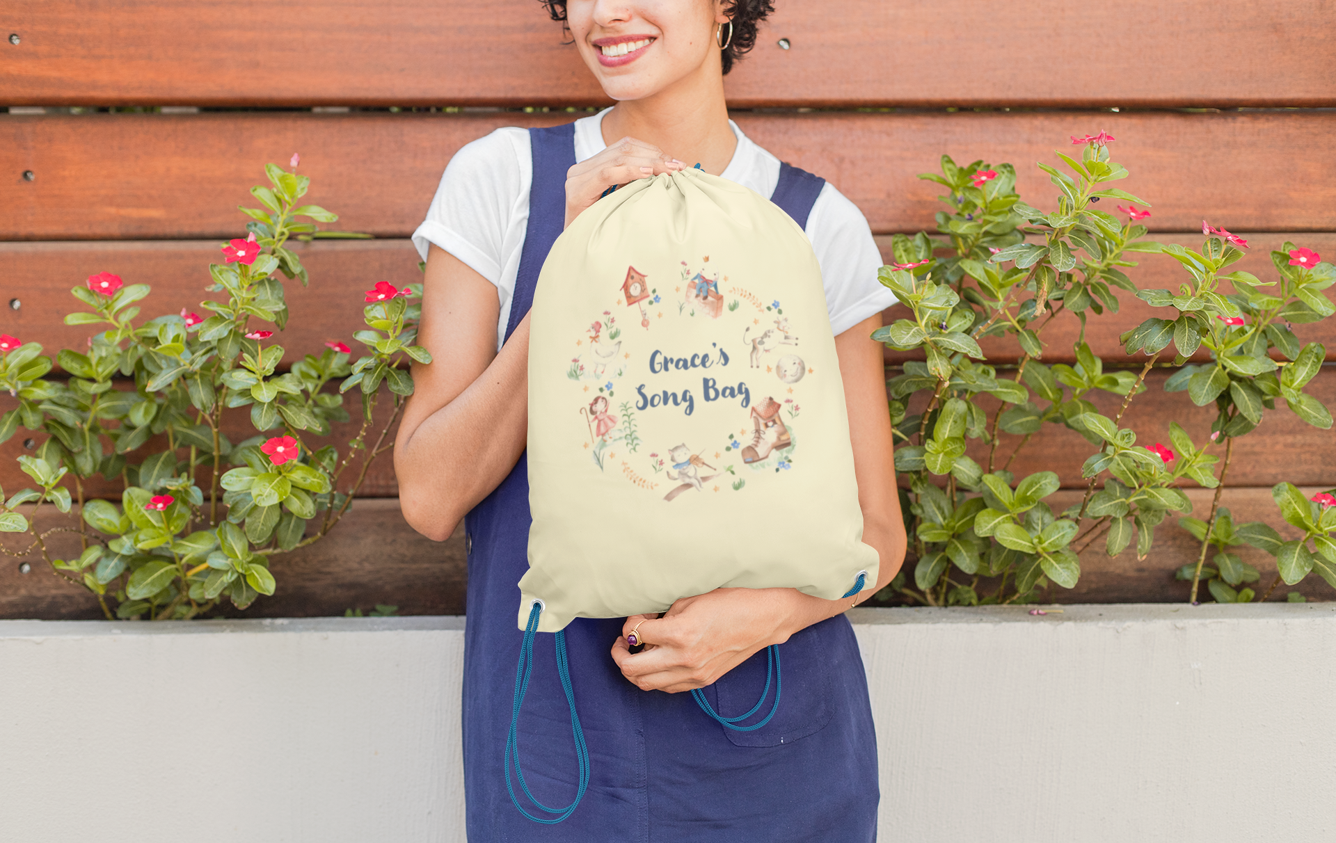 Image of woman holding a cream coloured cotton drawstring bag. Printed on the bag is some colourful illustrations from nursery rhymes, humpty dumpty, little bo peep, a clock, a boot, a goos, a cat and the fiddle and a cow and a moon. The watercolour illustrations are arranged in a wreath shape with the words 'Grace's son bang' written in blue in the middle.